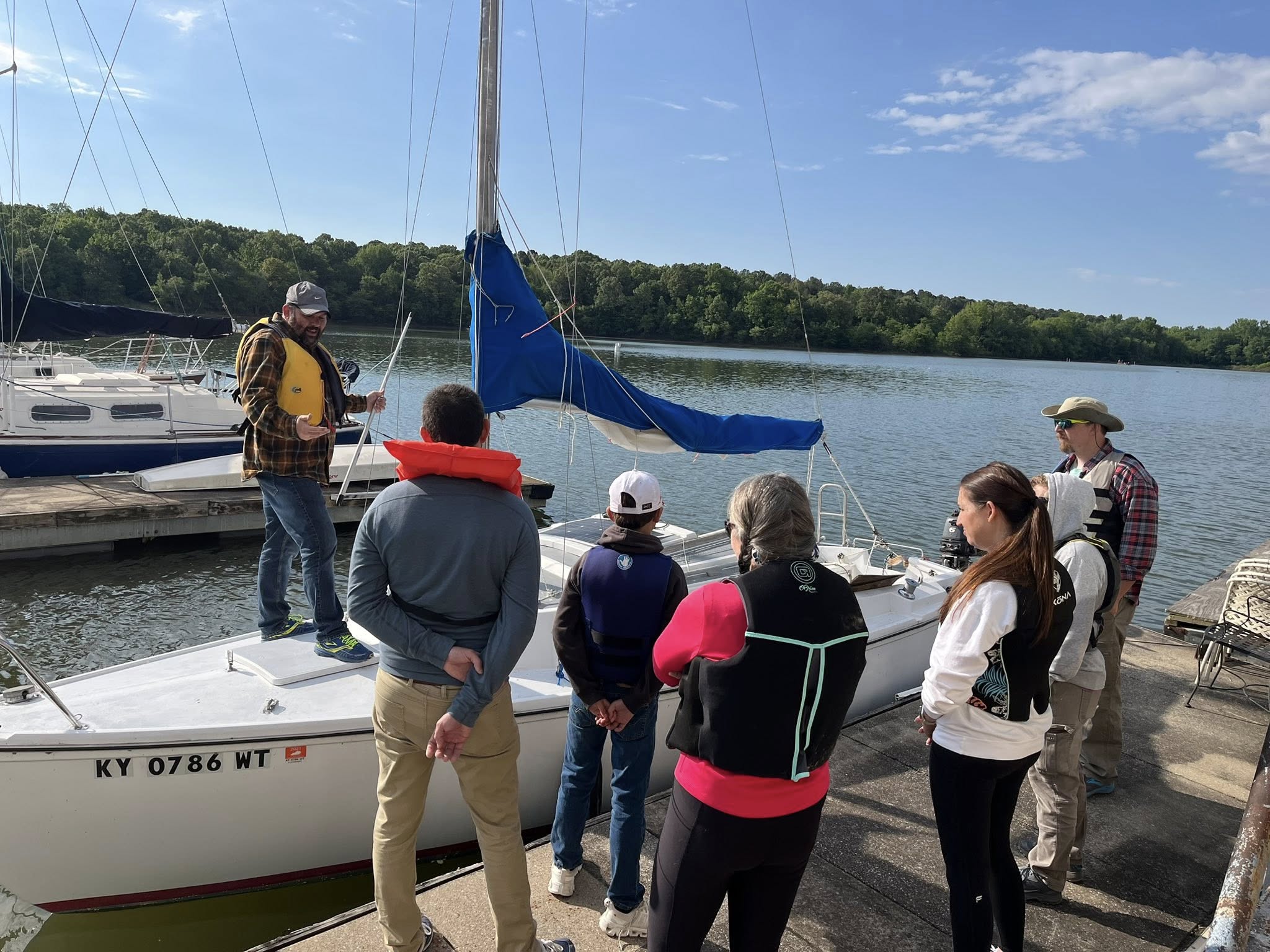 Richard Hall sailing club Richard Hall stands on a sailboat against a dock at Kentucky Dam Marina. He is speaking to a group of adults and kids, all students of Bluegrass Sailing Club.