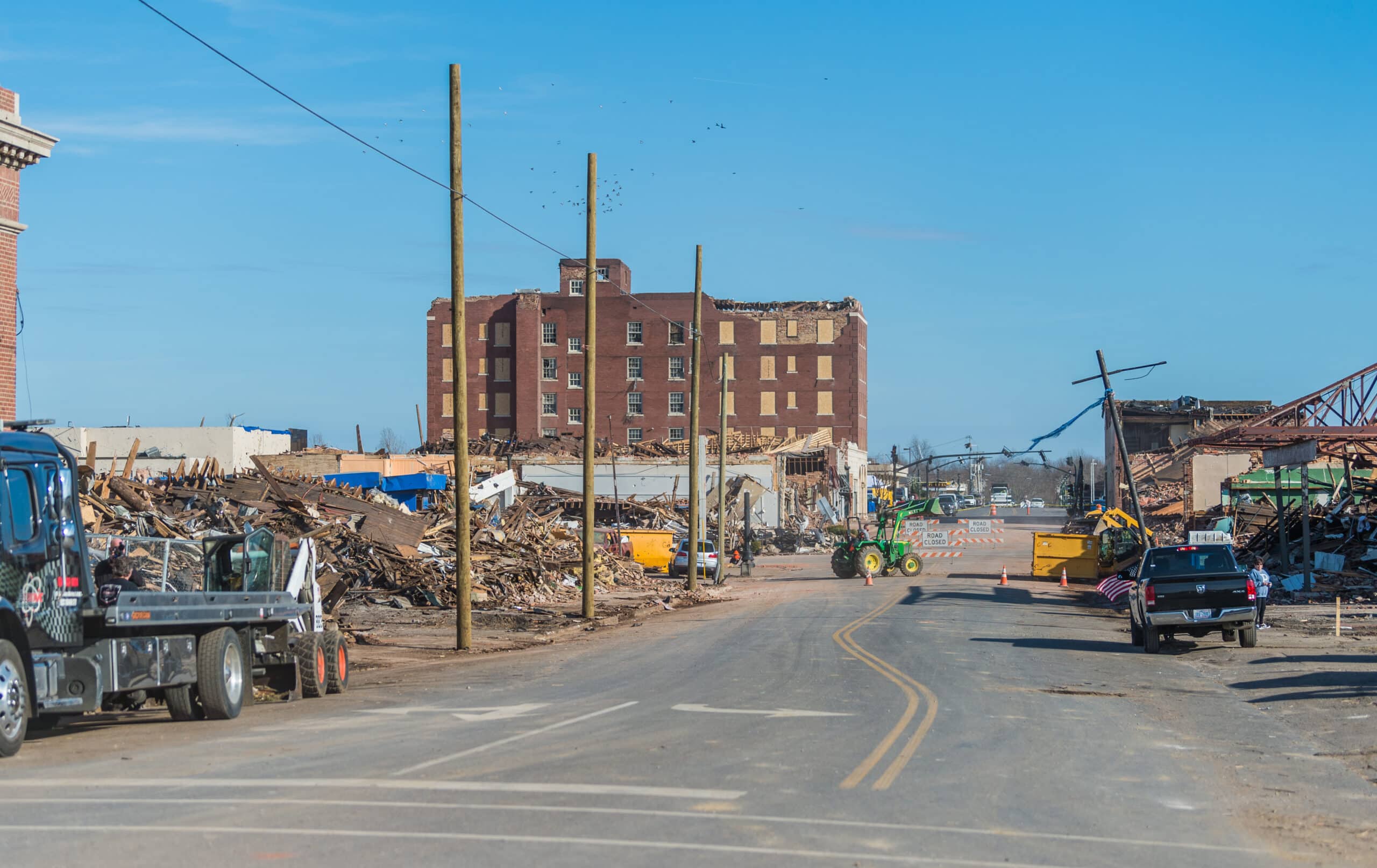 Downtown Mayfield, Kentucky, with damaged buildings, debris, work vehicles, and cleanup crews following the devastating December 2021 tornado.