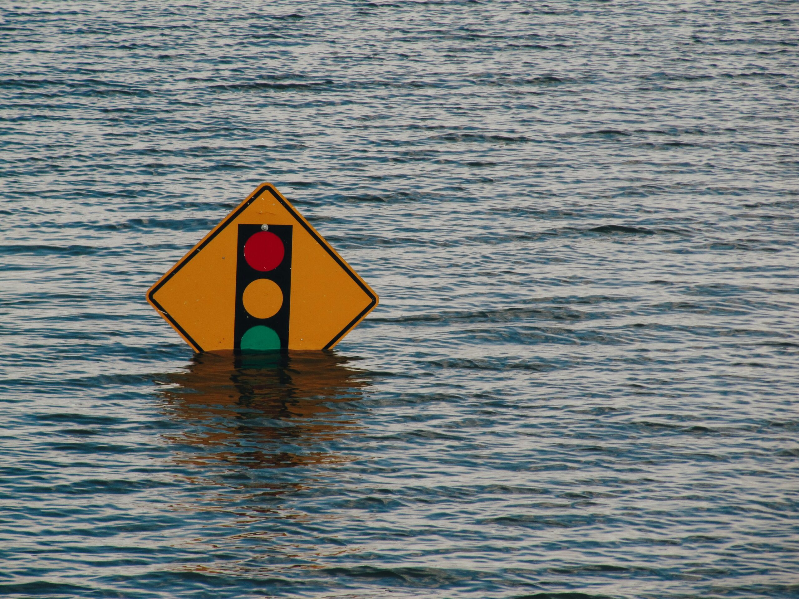 Traffic signal sign partially submerged in floodwater, with only the upper portion of the sign visible above the waterline.